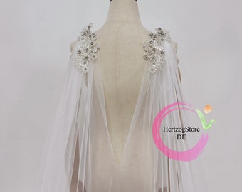 May include: White tulle bridal cape with beaded lace appliques on the shoulders. The cape is sheer and has a V-shaped back.