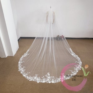 May include: A long, white bridal veil with a floral lace trim along the bottom edge. The veil is made of a sheer fabric and is displayed on a mannequin. A classic wedding accessory.