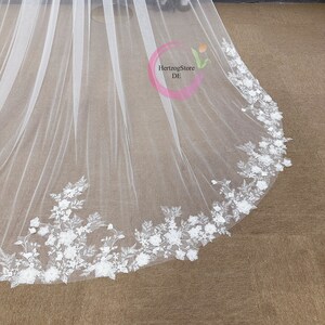 May include: White bridal veil with a delicate floral lace trim. The lace features white flowers and leaves.