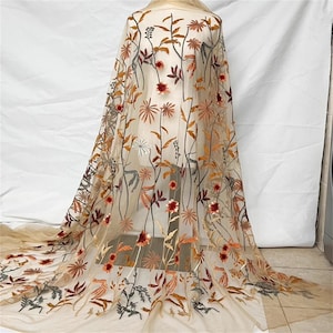 May include: Beige tulle fabric with embroidered floral design. The embroidery features flowers and leaves in shades of red, orange, brown, and green. The fabric is draped over a form, showcasing the intricate details of the design.