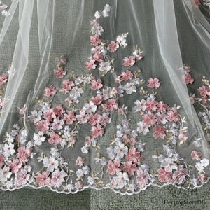 Pu&ograve; includere: Delicato tessuto di tulle bianco ornato da applicazioni floreali ricamate rosa e bianche. I fiori sono raggruppati lungo il bordo e sparsi sul materiale trasparente, creando un design romantico ed elegante. Il bordo smerlato aggiunge una finitura raffinata.