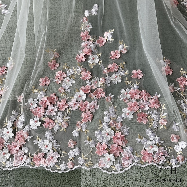 Pu&ograve; includere: Delicato tessuto di tulle bianco ornato da applicazioni floreali ricamate rosa e bianche. I fiori sono raggruppati lungo il bordo e sparsi sul materiale trasparente, creando un design romantico ed elegante. Il bordo smerlato aggiunge una finitura raffinata.