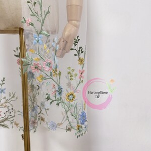 K&ouml;nnte beinhalten: Wei&szlig;er T&uuml;llstoff mit gestickten Blumenmotiven in Blaut&ouml;nen, Rosa, Gelb und Gr&uuml;n. Die Blumen sind &uuml;ber den Stoff verteilt und erzeugen einen zarten und romantischen Look.