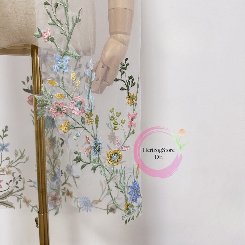 K&ouml;nnte beinhalten: Wei&szlig;er T&uuml;llstoff mit gestickten Blumenmotiven in Blaut&ouml;nen, Rosa, Gelb und Gr&uuml;n. Die Blumen sind &uuml;ber den Stoff verteilt und erzeugen einen zarten und romantischen Look.