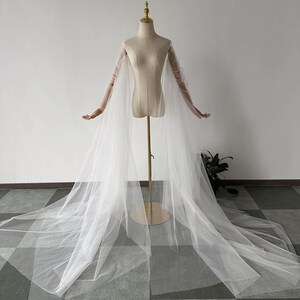 Könnte beinhalten: Ein weißer Braut-Umhang mit langem, fließendem Stoff und transparenten Ärmeln. Der Umhang wird auf einer Schaufensterpuppe mit goldenem Ständer präsentiert, um sein elegantes Design zu zeigen. Die transparenten Ärmel reichen bis zu den Händen der Schaufensterpuppe.
