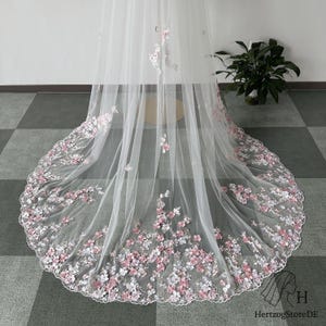 Pu&ograve; includere: Un velo da sposa bianco con delicati ricami floreali rosa e bianchi lungo l'orlo. Il velo &egrave; realizzato in tessuto trasparente e presenta un bordo smerlato, creando un look romantico ed elegante.