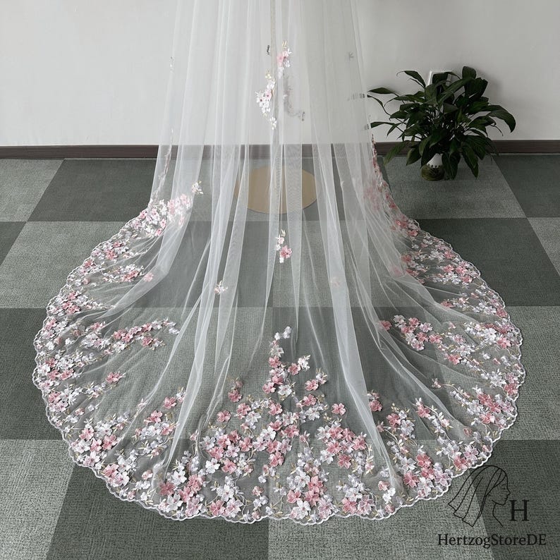 Pu&ograve; includere: Un velo da sposa bianco con delicati ricami floreali rosa e bianchi lungo l'orlo. Il velo &egrave; realizzato in tessuto trasparente e presenta un bordo smerlato, creando un look romantico ed elegante.