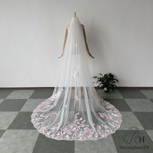 Pu&ograve; includere: Un lungo velo da sposa bianco con delicati ricami floreali rosa lungo il bordo inferiore e sparsi sul tulle. Il velo &egrave; esposto su un manichino, mostrando la sua lunghezza e il suo design. &Egrave; un accessorio da sposa.
