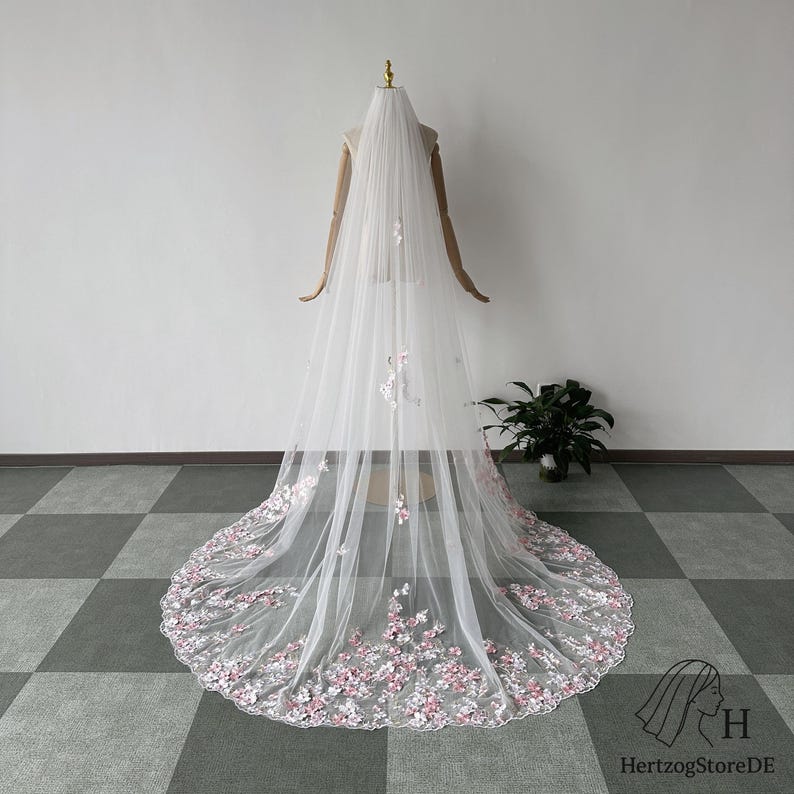 Pu&ograve; includere: Un lungo velo da sposa bianco con delicati ricami floreali rosa lungo il bordo inferiore e sparsi sul tulle. Il velo &egrave; esposto su un manichino, mostrando la sua lunghezza e il suo design. &Egrave; un accessorio da sposa.