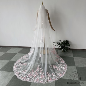 Pu&ograve; includere: Un lungo velo da sposa bianco con delicati ricami floreali nei toni del rosa e del bianco. Il velo &egrave; esposto su un manichino, mostrando la sua lunghezza e i suoi dettagli intricati. Il design floreale &egrave; concentrato lungo il bordo inferiore.