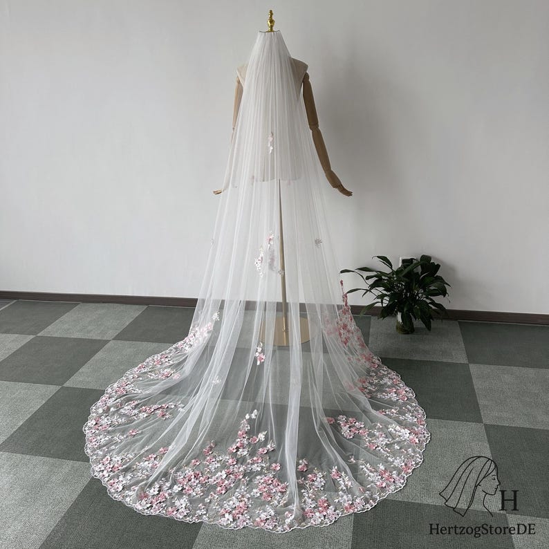 Pu&ograve; includere: Un lungo velo da sposa bianco con delicati ricami floreali nei toni del rosa e del bianco. Il velo &egrave; esposto su un manichino, mostrando la sua lunghezza e i suoi dettagli intricati. Il design floreale &egrave; concentrato lungo il bordo inferiore.