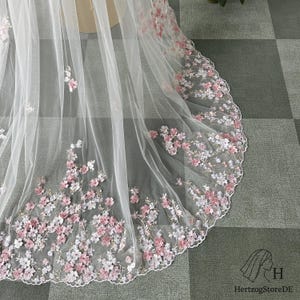 Pu&ograve; includere: Un velo da sposa bianco trasparente con bordo smerlato, ornato da delicati ricami floreali rosa e bianchi. Il velo &egrave; realizzato in un tessuto leggero e fluido, perfetto per un matrimonio.