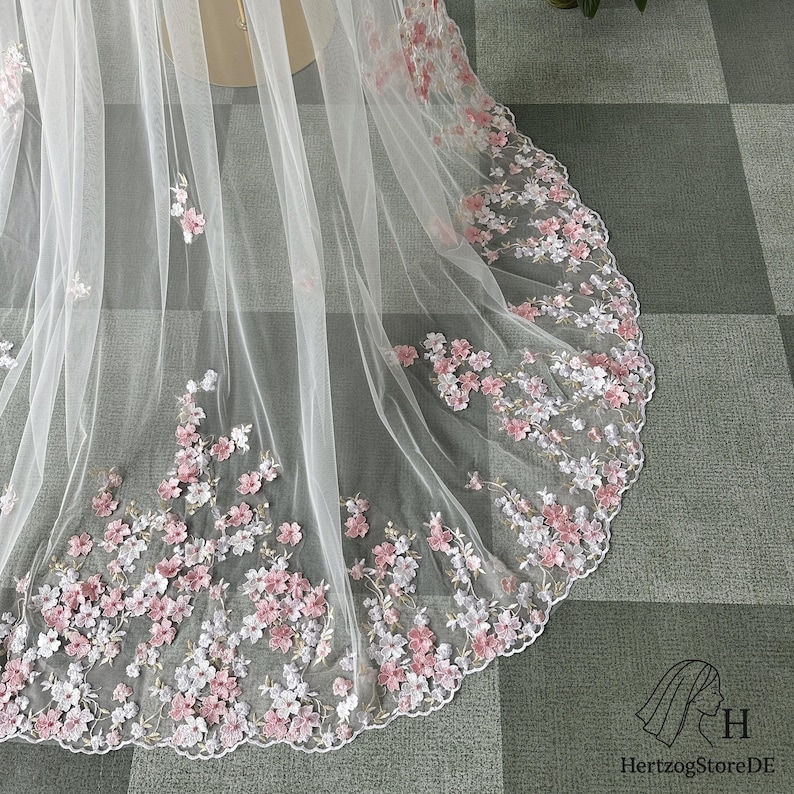 Pu&ograve; includere: Un velo da sposa bianco trasparente con bordo smerlato, ornato da delicati ricami floreali rosa e bianchi. Il velo &egrave; realizzato in un tessuto leggero e fluido, perfetto per un matrimonio.