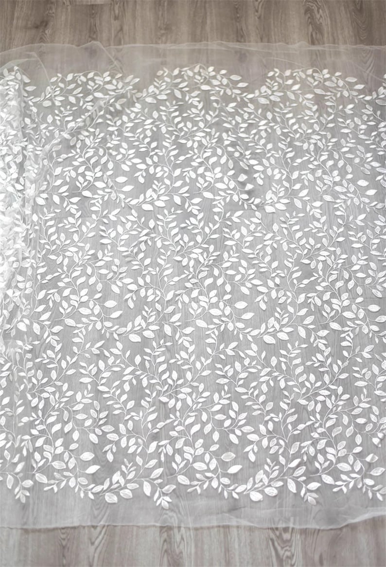 May include: White embroidered fabric with a delicate leaf pattern. The fabric is sheer and has a soft, flowing texture.