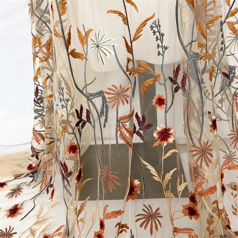 May include: Sheer fabric with embroidered floral design. The embroidery features a variety of flowers and leaves in shades of orange, brown, red, and gray. The fabric is a light beige color, and the floral design is intricately detailed.