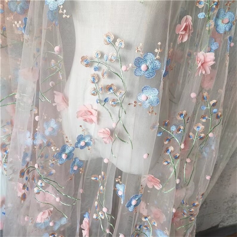 May include: Pale pink tulle fabric with embroidered blue and pink flowers. The fabric has a delicate, feminine look and would be perfect for a variety of projects.