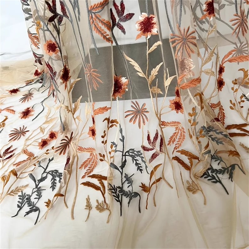 May include: Sheer beige curtain with embroidered floral design. The embroidery features flowers and leaves in shades of burgundy, orange, brown, and gray. The curtain is made of a lightweight, transparent fabric, allowing light to pass through.