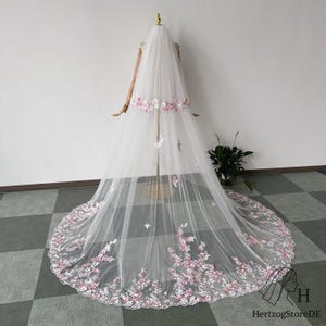 May include: A long, white bridal veil with delicate pink floral embroidery. The veil is displayed on a mannequin, showcasing the intricate details of the flowers and the flowing fabric. The floral design is concentrated at the edges and along the top.
