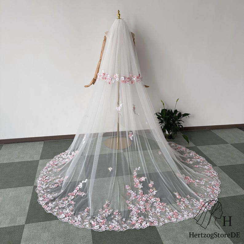 May include: A long, white bridal veil with delicate pink floral embroidery. The veil is displayed on a mannequin, showcasing the intricate details of the flowers and the flowing fabric. The floral design is concentrated at the edges and along the top.
