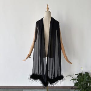 Op de afbeelding: Een zwarte, doorschijnende cape met een veren rand, tentoongesteld op een mannequin. De cape is over de schouders gedrapeerd en hangt naar voren. De veren rand zit aan de onderkant van de cape.