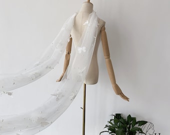 May include: A white bridal veil with butterfly and pearl embellishments is draped over a mannequin. The veil is made of a sheer fabric and features delicate butterfly appliqués and scattered pearls. The mannequin is on a gold stand.