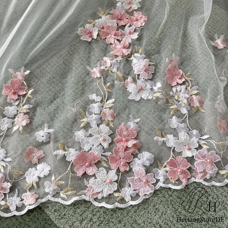 May include: Delicate floral lace fabric featuring embroidered pink and white flowers with gold accents. The flowers are clustered along the edge and scattered across the sheer, off-white material. The lace has a scalloped edge.