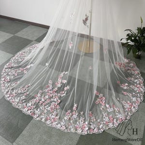 May include: A white bridal veil with delicate pink and white floral embroidery along the edge. The veil is made of sheer fabric and is displayed on a gold stand. The floral design adds a touch of elegance.