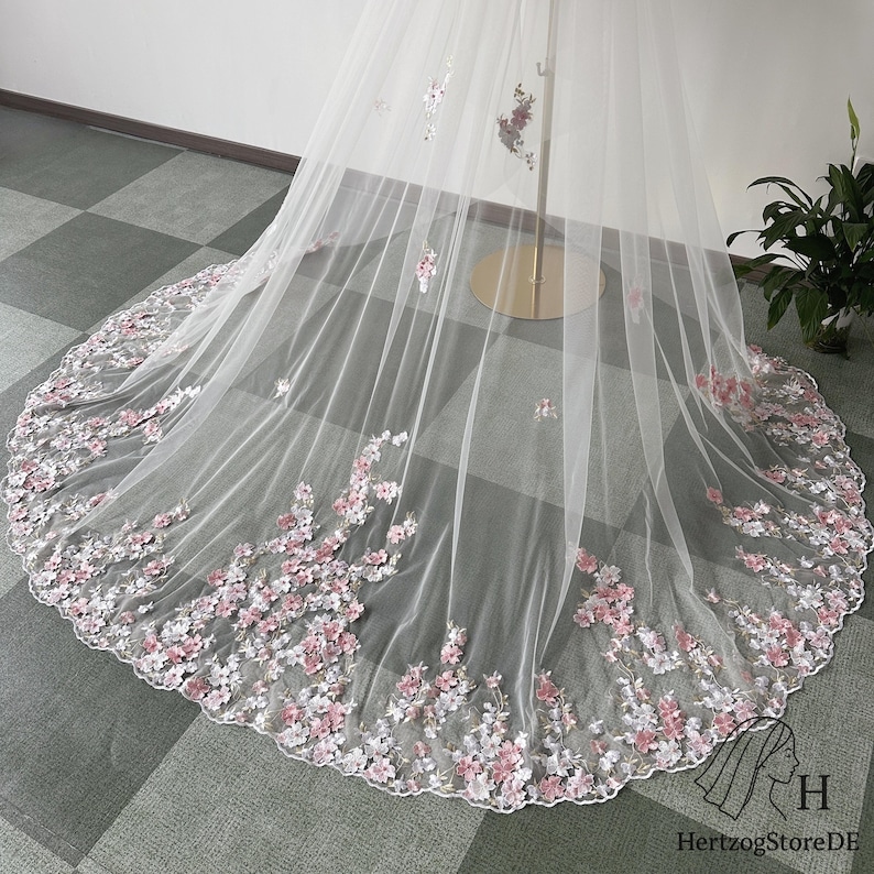May include: A white bridal veil with delicate pink and white floral embroidery along the edge. The veil is made of sheer fabric and is displayed on a gold stand. The floral design adds a touch of elegance.