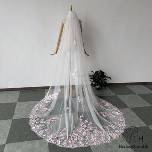 Pu&ograve; includere: Un lungo velo da sposa bianco con delicati ricami floreali rosa e bianchi lungo l'orlo. Il velo &egrave; esposto su un manichino, mostrando la sua lunghezza e il suo design intricato. Il tessuto trasparente e i dettagli floreali creano un'estetica romantica.