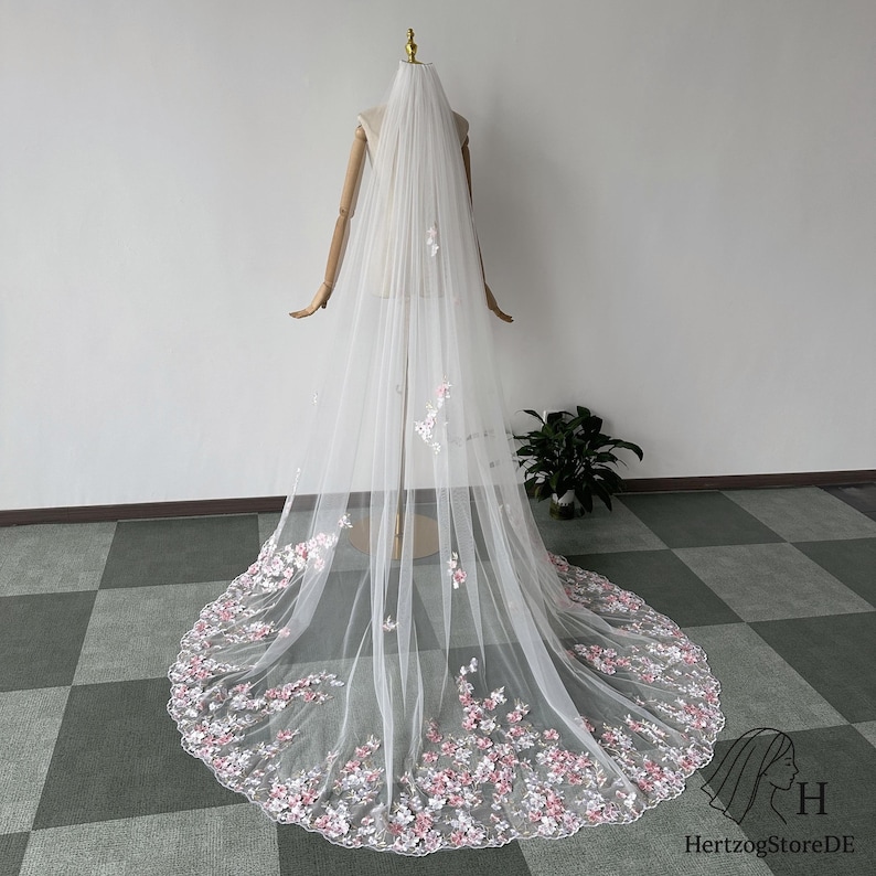 Pu&ograve; includere: Un lungo velo da sposa bianco con delicati ricami floreali rosa e bianchi lungo l'orlo. Il velo &egrave; esposto su un manichino, mostrando la sua lunghezza e il suo design intricato. Il tessuto trasparente e i dettagli floreali creano un'estetica romantica.
