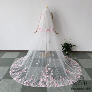 May include: A long, white bridal veil with delicate pink and white floral embroidery. The veil is displayed on a mannequin, showcasing the intricate floral design along the edges and at the top, creating a romantic and elegant look.