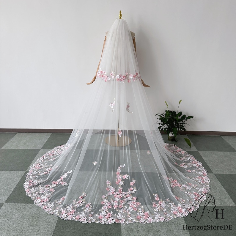 May include: A long, white bridal veil with delicate pink and white floral embroidery. The veil is displayed on a mannequin, showcasing the intricate floral design along the edges and at the top, creating a romantic and elegant look.