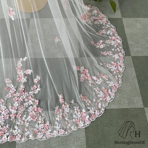 May include: A sheer white veil with delicate floral embroidery in shades of pink and white. The floral design is concentrated along the scalloped edge, creating a romantic and elegant look. The veil is likely for a wedding or special occasion.