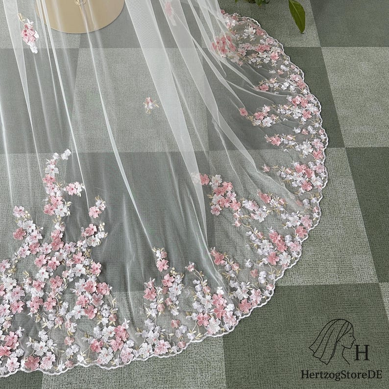 May include: A sheer white veil with delicate floral embroidery in shades of pink and white. The floral design is concentrated along the scalloped edge, creating a romantic and elegant look. The veil is likely for a wedding or special occasion.