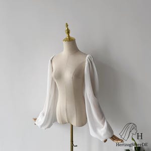以下が含まれることがあります： 白い、シアーなパフスリーブの長袖ブラウスがマネキンに展示されています。ブラウスはシンプルでエレガントなデザインで、フォーマルにもカジュアルにも最適です。マネキンは金色のスタンドに立っています。
