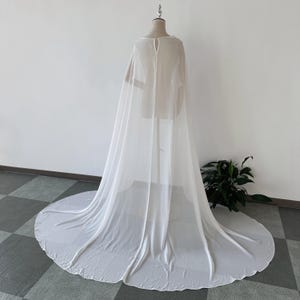 K&ouml;nnte beinhalten: Ein wei&szlig;er, durchsichtiger Chiffon-Umhang mit langem Schleier. Der Umhang ist &uuml;ber eine Schaufensterpuppe gelegt und hat ein schlichtes, elegantes Design.