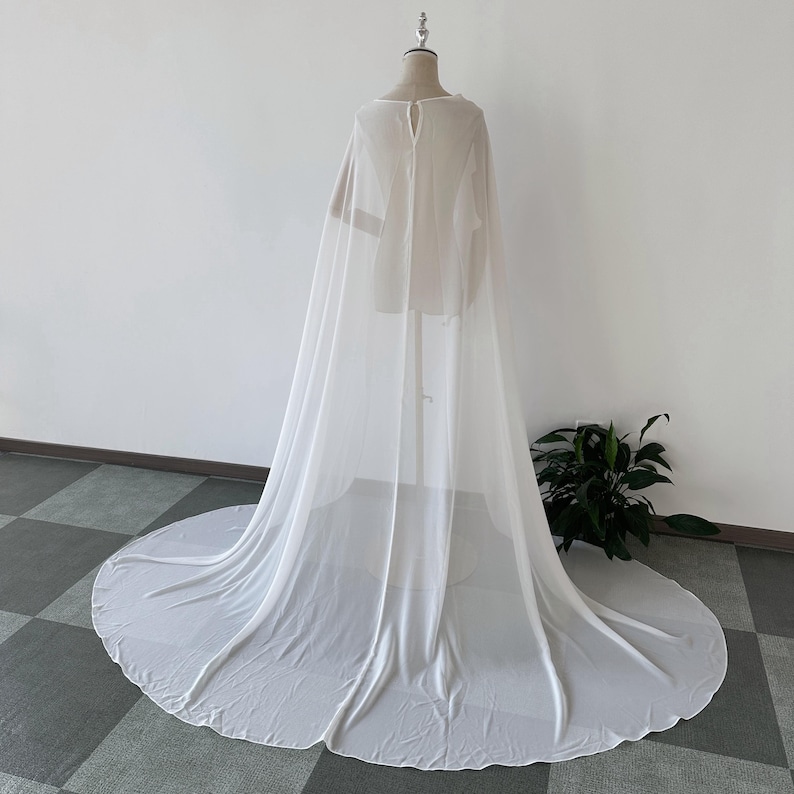 K&ouml;nnte beinhalten: Ein wei&szlig;er, durchsichtiger Chiffon-Umhang mit langem Schleier. Der Umhang ist &uuml;ber eine Schaufensterpuppe gelegt und hat ein schlichtes, elegantes Design.