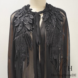 May include: A sheer black cape featuring large, detailed black embroidered wing appliques on the front shoulders. The garment includes a tie closure at the neckline and is displayed on a mannequin. The logo HertzogStoreDE is visible.