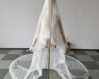 May include: A white bridal veil with delicate lace trim, displayed on a mannequin. The veil features multiple layers and a long train, creating an elegant and romantic look. The lace detailing adds a touch of sophistication to the design.