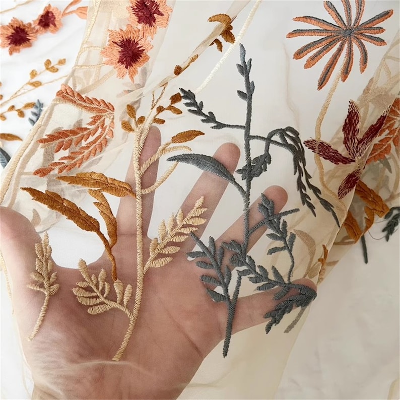 May include: Close-up of sheer fabric with embroidered floral and botanical designs. The embroidery features orange, rust, and gray colors. The fabric is held in a hand, showcasing the delicate details of the needlework.