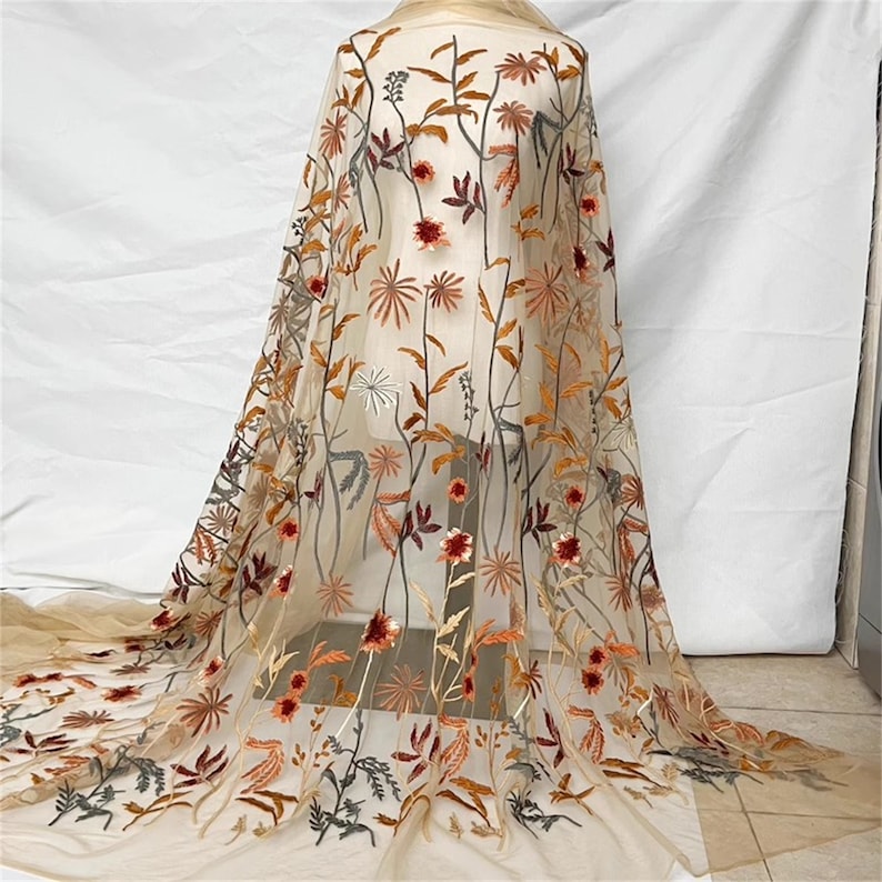 May include: Beige tulle fabric with embroidered floral design. The embroidery features flowers and leaves in shades of red, orange, brown, and green. The fabric is draped over a form, showcasing the intricate details of the design.
