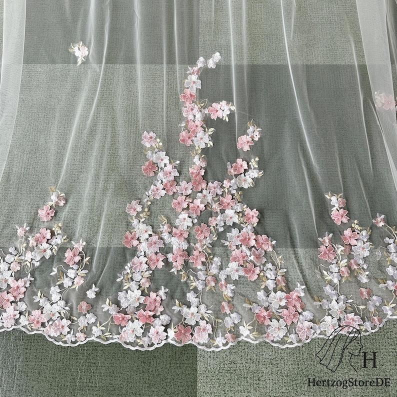 May include: Sheer white fabric with embroidered pink and white floral design. The delicate flowers are clustered along the bottom edge and scattered across the fabric. The fabric has a scalloped edge.