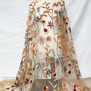 May include: Sheer fabric with an embroidered floral design. The embroidery features flowers and leaves in shades of orange, red, and brown, set against a beige background. The fabric is draped, showcasing the intricate details of the floral pattern.