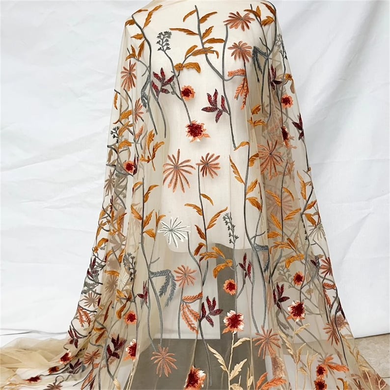 May include: Sheer fabric with an embroidered floral design. The embroidery features flowers and leaves in shades of orange, red, and brown, set against a beige background. The fabric is draped, showcasing the intricate details of the floral pattern.