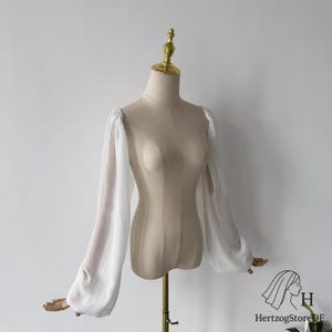 以下が含まれることがあります： マネキンに展示された、白いシースルー袖の衣服。袖は長くボリュームがあり、肩と手首にギャザーのディテールがあります。マネキンはベージュ色で、背景は無地の白です。