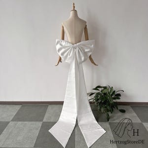 Peut inclure: Un grand nœud en satin blanc avec de longues queues flottantes, présenté sur un mannequin. Le nœud est le point central, avec un design plein et volumineux. Les queues s'étendent vers le bas, créant un effet dramatique. Le fond est blanc neutre.
