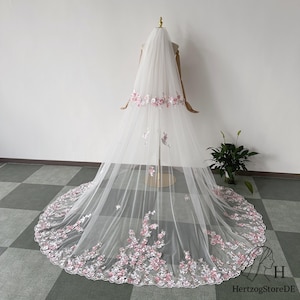 May include: A long, white bridal veil with delicate pink floral embroidery along the edges and scattered across the tulle. The veil is displayed on a mannequin, showcasing its flowing design. The floral details add a touch of elegance.