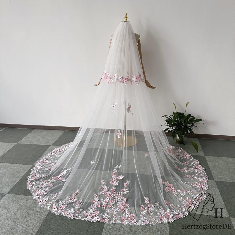 May include: A long, white bridal veil with delicate pink floral embroidery along the edges and scattered across the tulle. The veil is displayed on a mannequin, showcasing its flowing design. The floral details add a touch of elegance.