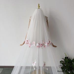 May include: A white bridal veil with delicate pink and white floral embroidery along the bottom edge. The veil is made of a sheer, flowing fabric and is displayed on a mannequin.