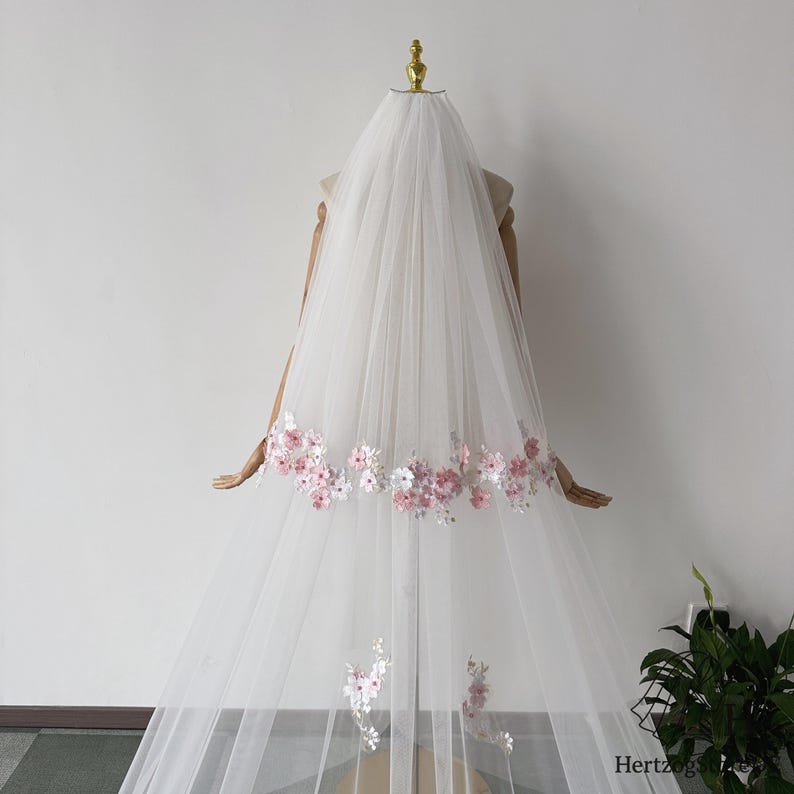 May include: A white bridal veil with delicate pink and white floral embroidery along the bottom edge. The veil is made of a sheer, flowing fabric and is displayed on a mannequin.